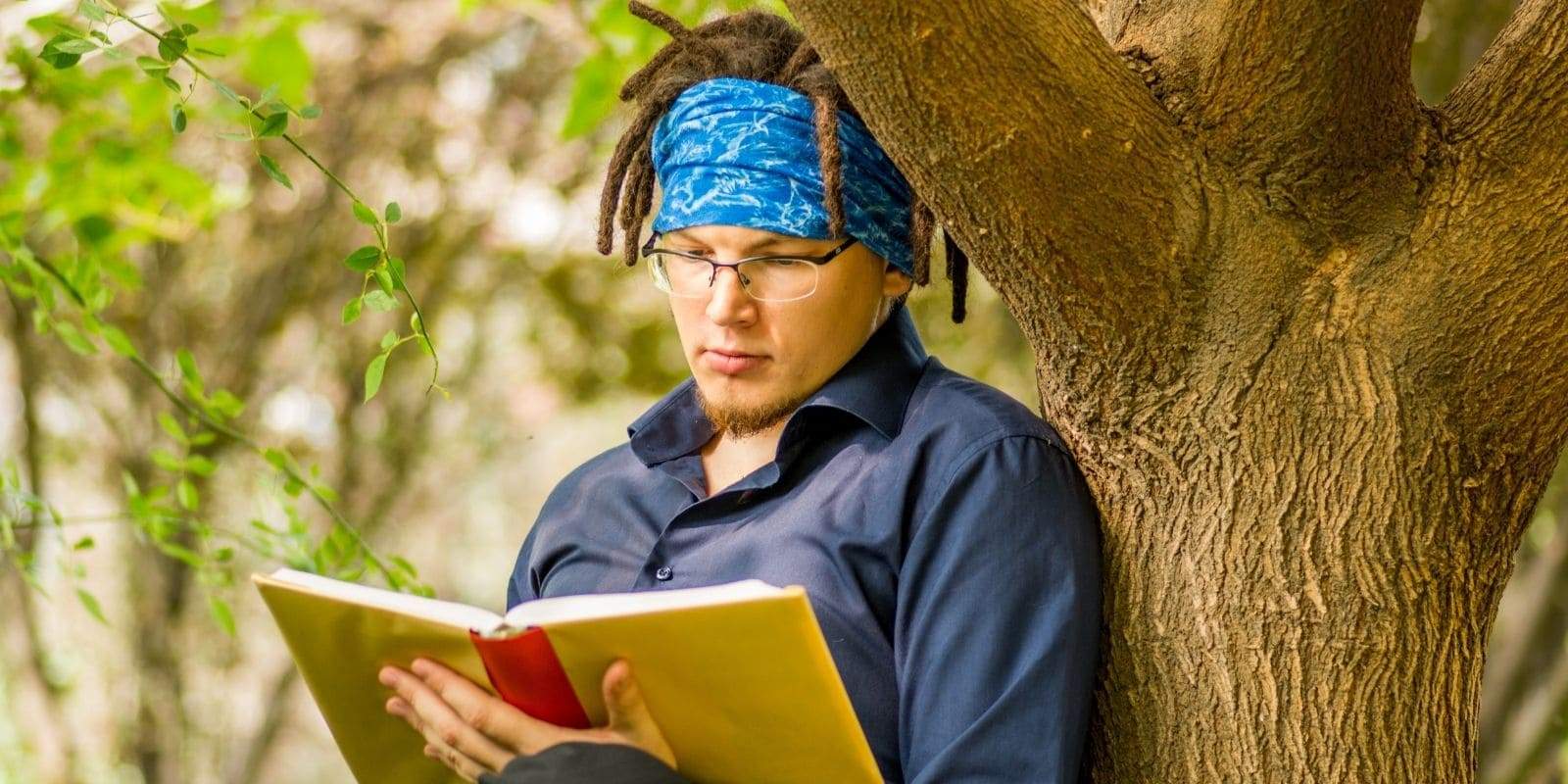 Homme avec dreadlocks pose contre un arbre en train de lire